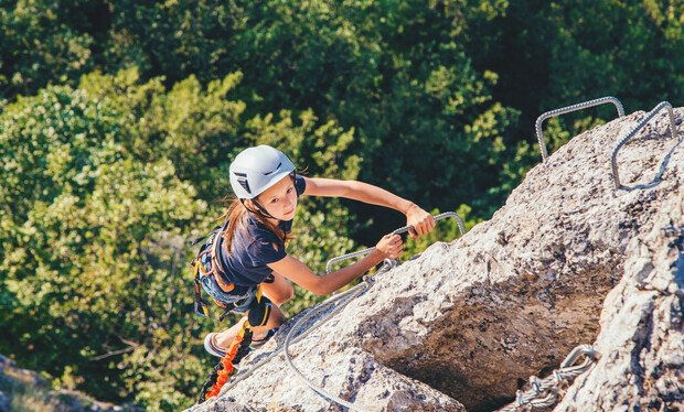 A climber wearing a helmet and harness ascends a rocky cliff using metal rungs, with lush green foliage visible in the background.