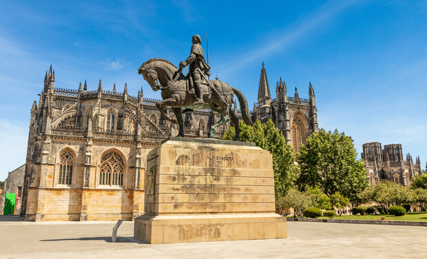 A bronze equestrian statue stands on a stone pedestal inscribed “DOM NUNES ALVARES PEREIRA” before a large, ornate Gothic cathedral, bordered by trees and set against a clear blue sky.