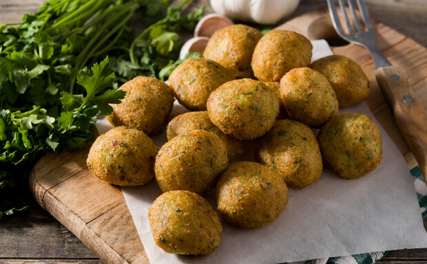 Falafel balls rest on parchment paper atop a wooden cutting board, surrounded by fresh parsley, garlic, and utensils in a rustic kitchen setting.