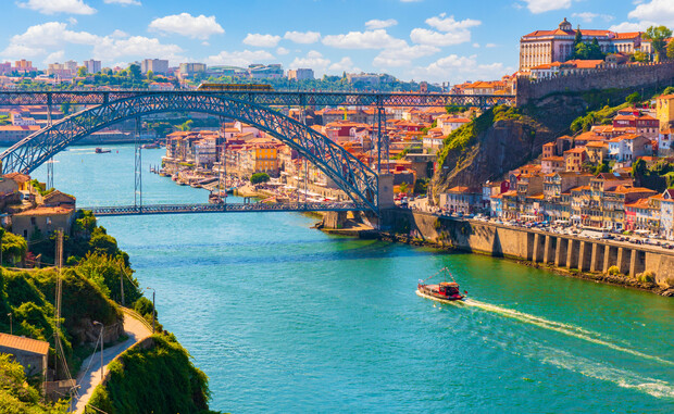 A boat travels under a large, arched metal bridge over a river. Colorful buildings line the riverbanks, with a hilltop structure visible against a partly cloudy sky.
