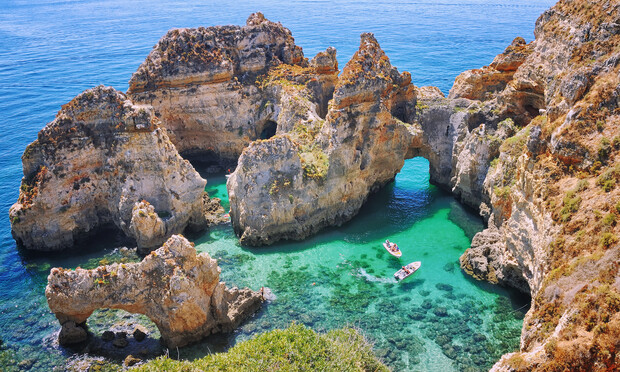 Rock formations create a stunning archway over clear turquoise waters; boats float below in a serene coastal landscape, surrounded by rugged cliffs under a bright blue sky.
