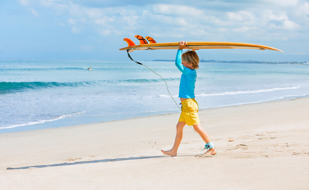 A child carries a surfboard overhead while walking along a sandy beach. The ocean waves softly crash in the background under a cloudy sky.