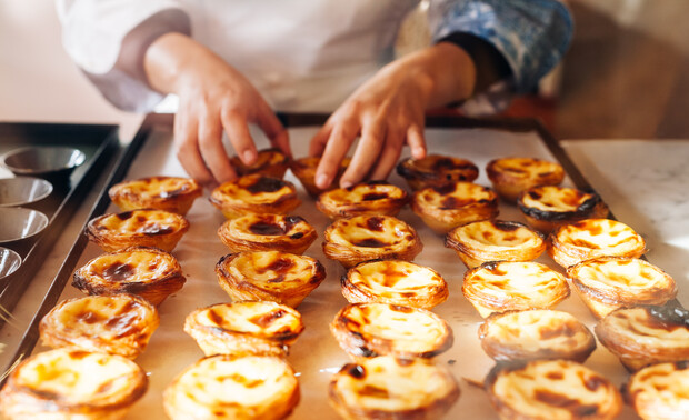 Hands arrange freshly baked custard tarts on a baking tray, surrounded by a warm kitchen environment with soft lighting highlighting the pastries' golden crusts.
