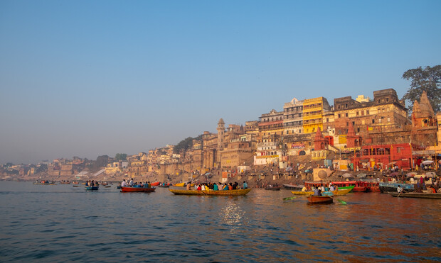 Boats float on a river beside a densely packed row of colorful, historic buildings under a clear blue sky, creating a vibrant and bustling waterfront scene.