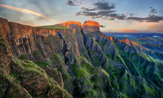 Majestic cliffs bask in the warm glow of a setting sun, with steep rock faces contrasting lush green valleys; clouds scatter across a vibrant sky, enhancing the serene landscape.
