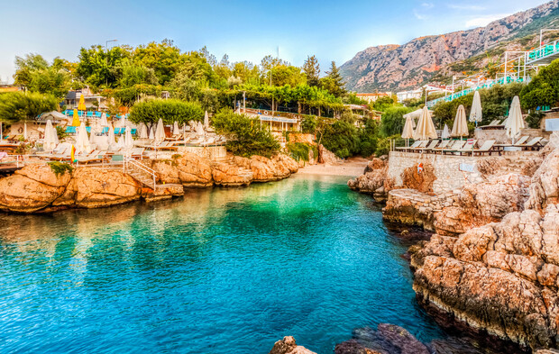 Rocky bay with turquoise water, bordered by sun loungers and umbrellas. Trees cover the cliffs, with mountains visible in the background under a clear sky.