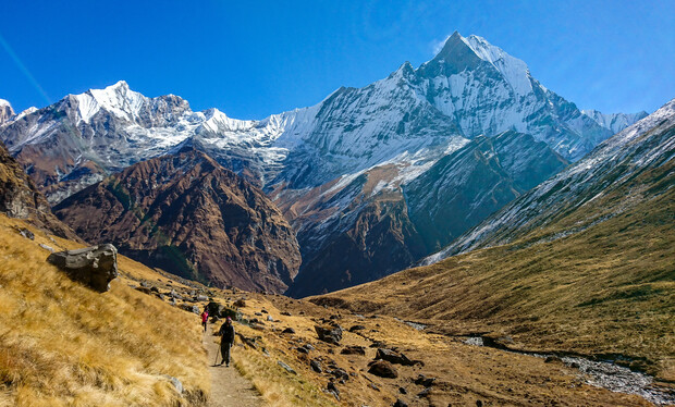 Hikers trek along a narrow path in a mountainous landscape, with snow-capped peaks and rugged terrain under a clear blue sky, evoking a sense of adventure and natural beauty.