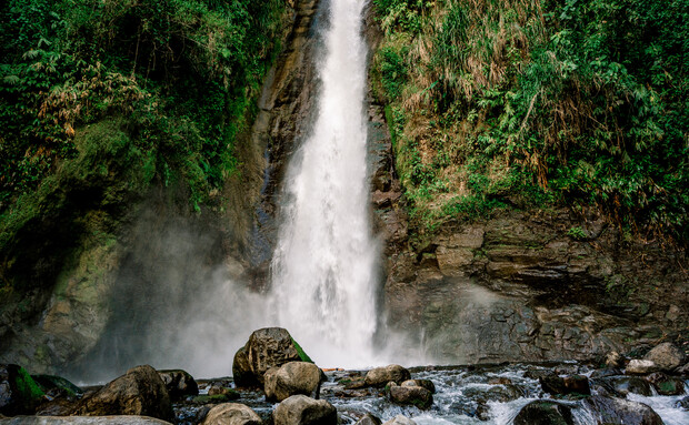 A waterfall cascades down a rocky cliff, surrounded by lush greenery. Water crashes into a pool below, creating mist and flowing over scattered rocks in a forested setting.