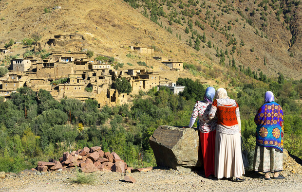 Three women in colorful clothing stand together, looking towards a hillside village consisting of mud-brick houses, surrounded by sparse vegetation under clear blue skies.