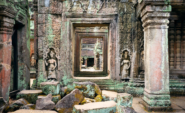 Stone doorways with intricate carvings line up in succession through an ancient temple, flanked by statues of figures, surrounded by fallen rocks and aged, weathered walls.