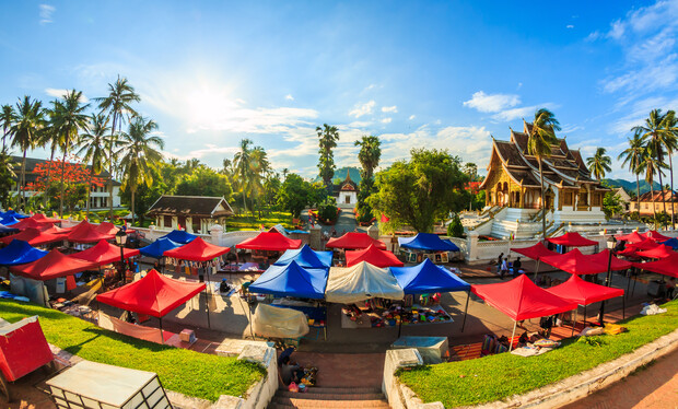 Colorful market stalls with red and blue canopies sit under a sunny sky. A historic temple building is visible in the background, surrounded by lush greenery and palm trees.