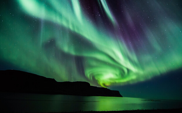 Aurora borealis swirls across the night sky, illuminating a coastal landscape with vivid greens and purples, reflecting on calm water below.