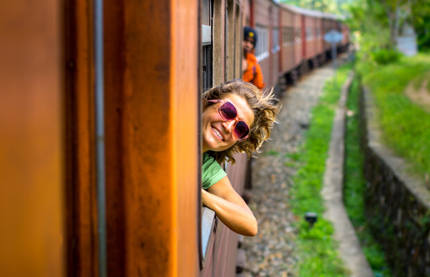 A person in sunglasses leans out a moving train window, smiling. The train travels on tracks through a lush, green landscape, with another person visible in the distance.