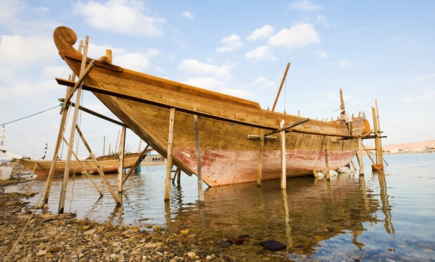 A wooden boat under construction stands on supports, partially submerged in shallow, rocky waters, with a clear blue sky and a distant shoreline in the background.