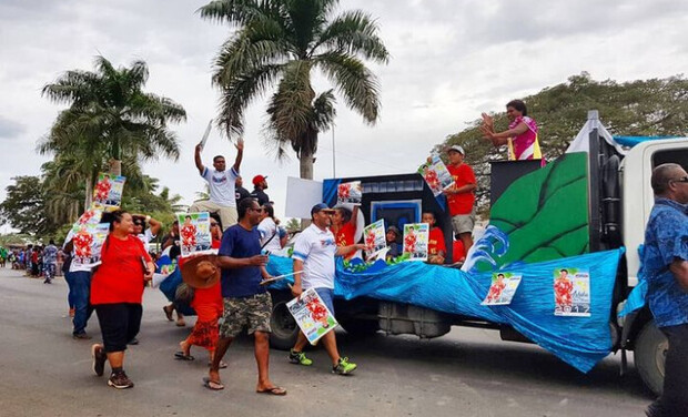 A group of people on a decorated truck and walking beside it, carrying colorful posters. They are participating in a parade on a palm-lined street, under a cloudy sky.