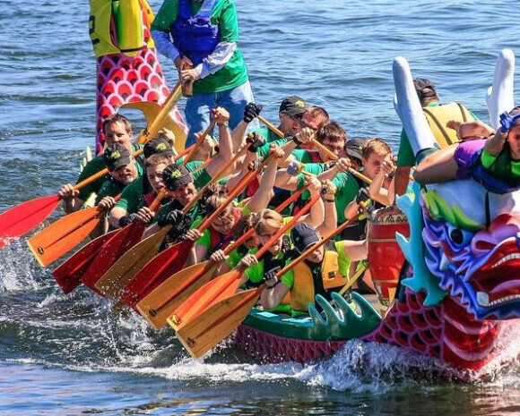 Dragon boat team paddles vigorously, splashing water, in a colorful dragon-shaped boat. Participants wear life vests and paddle in unison. The setting is a sunny day on a calm body of water.
