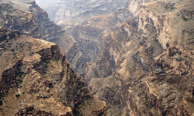Rocky cliffs form a deep canyon, showcasing steep, rugged walls. Sparse vegetation dots the barren landscape under a hazy sky, indicating a remote, arid environment.