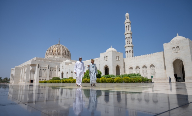 Two individuals walk across a reflective courtyard in front of a large, ornate mosque featuring a prominent dome and minaret, under a clear blue sky.
