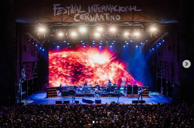 Band performing energetically on an illuminated stage with a fiery cosmic background screen, surrounded by a large, cheering crowd. Overhead, text reads "FESTIVAL INTERNACIONAL CERVANTINO."