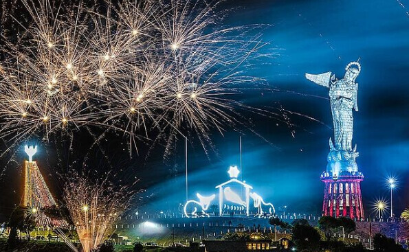 Statue of a figure with open arms is illuminated in blue and red light. Fireworks explode overhead. Nearby, a structure decorated with festive lights and a glowing star adds visual interest.