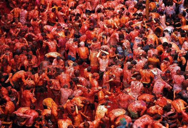 People covered in tomatoes engage in a massive food fight, throwing and splashing each other in a crowded outdoor festival, surrounded by a sea of red.