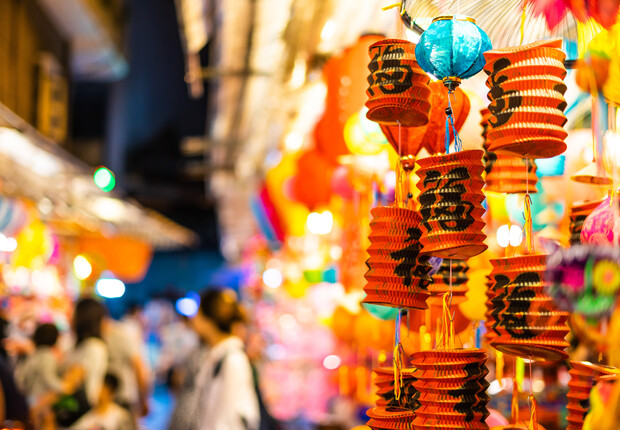 Colorful paper lanterns hang in a bustling street market, glowing warmly. Blurred people move around, creating a vibrant, festive atmosphere. Chinese characters are visible on some lanterns.