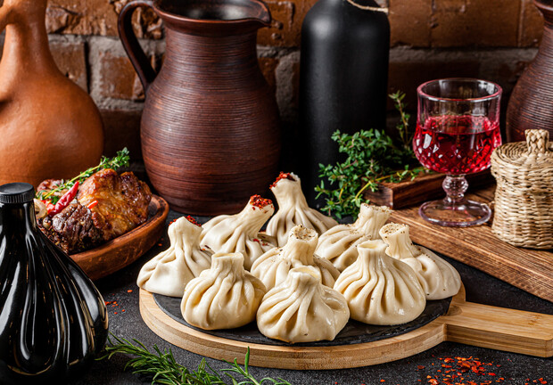Dumplings rest on a wooden board garnished with herbs, surrounded by ceramic jugs, a plate of roasted meat, and a glass of red wine on a wooden table, brick wall backdrop.