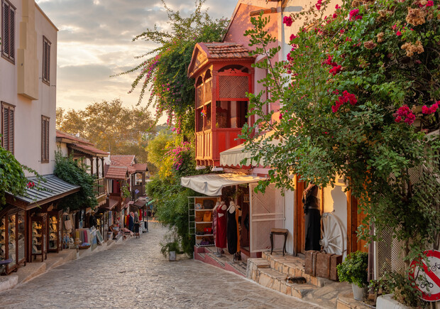 A cobblestone street features shops with colorful displays and textiles, bordered by lush greenery and flowers, under a partially cloudy sky with warm, early evening lighting.