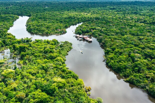 A river winds through lush green rainforest. A boat creates a wake as it moves along the water towards a cluster of small buildings on the riverbank.