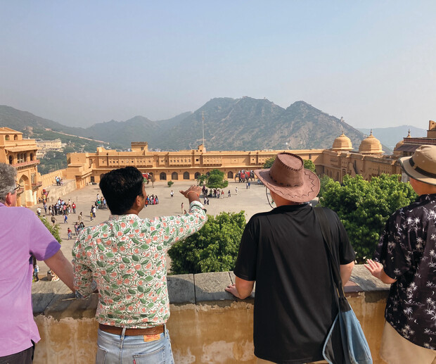 Four people are standing on a balcony overlooking a square at a historic fort, with mountains visible in the background. One individual points towards the scene, possibly explaining something.