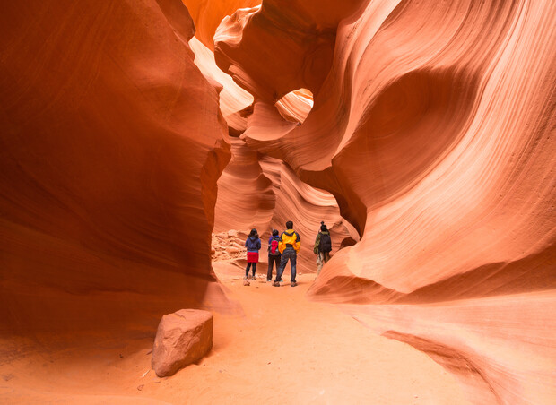 People stand inside a winding, narrow sandstone canyon, likely in a desert. The rock walls exhibit smooth, flowing patterns in warm shades of orange and red, with sunlight illuminating the scene.
