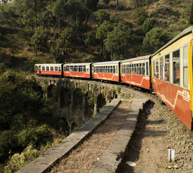 A red and cream train traverses a curving stone bridge, surrounded by trees covering a hilly landscape. The train is marked with the number "416".