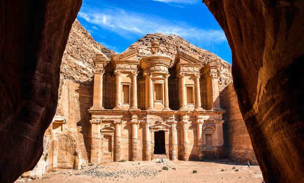 Ancient carved stone façade stands majestically, framed by rocky cliffs, under a clear blue sky. The surrounding desert terrain enhances the monumental structure's grandeur.