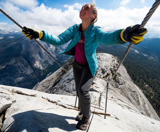 A person climbs a steep rock face using metal cables, wearing gloves and a blue jacket, surrounded by rugged mountain scenery under a partly cloudy sky.