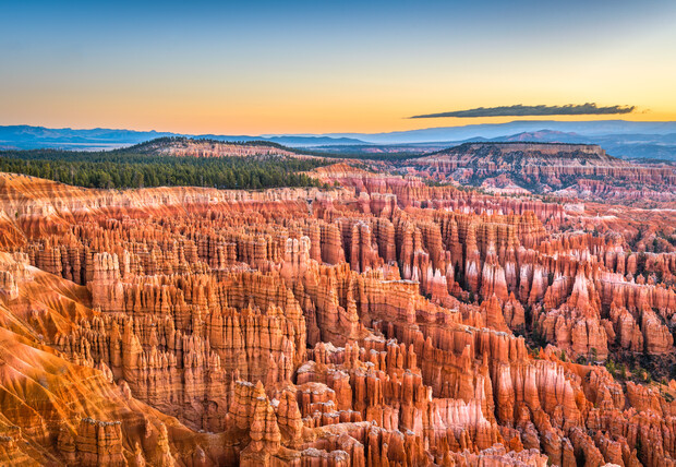 Eroded rock formations rise dramatically, illuminated by sunset hues, creating a vast, rugged landscape. In the distance, forested plateaus stretch beneath a clear sky, with a lone cloud overhead.