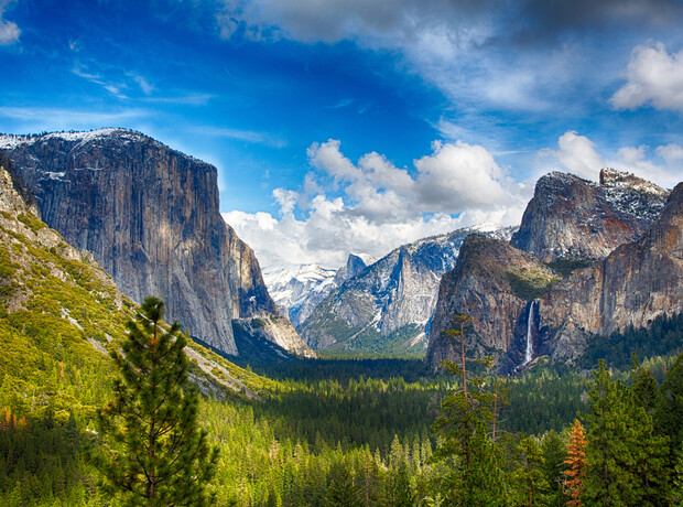 Mountains stretch across the landscape, with a waterfall cascading down rocky cliffs, surrounded by lush green forests and under a vibrant blue sky with scattered clouds.