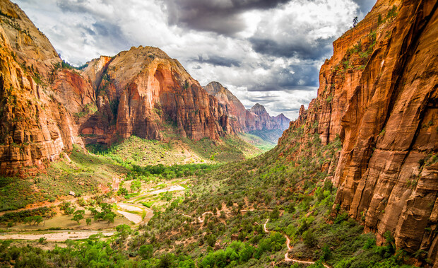 Canyon walls tower over a winding valley, where lush greenery contrasts against red rock formations under a dramatic, cloudy sky.