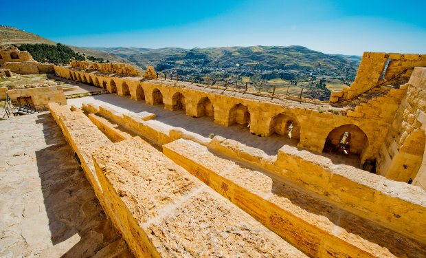 Ancient stone fortress with arches stands under a clear blue sky, overlooking a vast landscape of rolling hills and valleys.