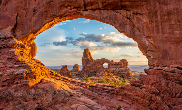 A natural sandstone arch frames a desert landscape, with rocky formations and a partly cloudy sky, under golden sunset light in a vast, arid environment.