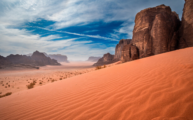 Sweeping sand dunes stretch across the desert, bordered by towering rocky cliffs under a bright blue sky with scattered clouds. Distant mountains fade into the horizon, completing the arid landscape.