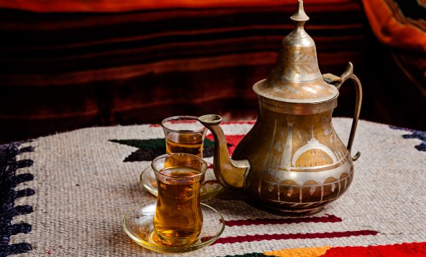 A decorative metal teapot and two small glasses filled with tea rest on a woven, multicolored textile, possibly within a cozy, dimly lit room.