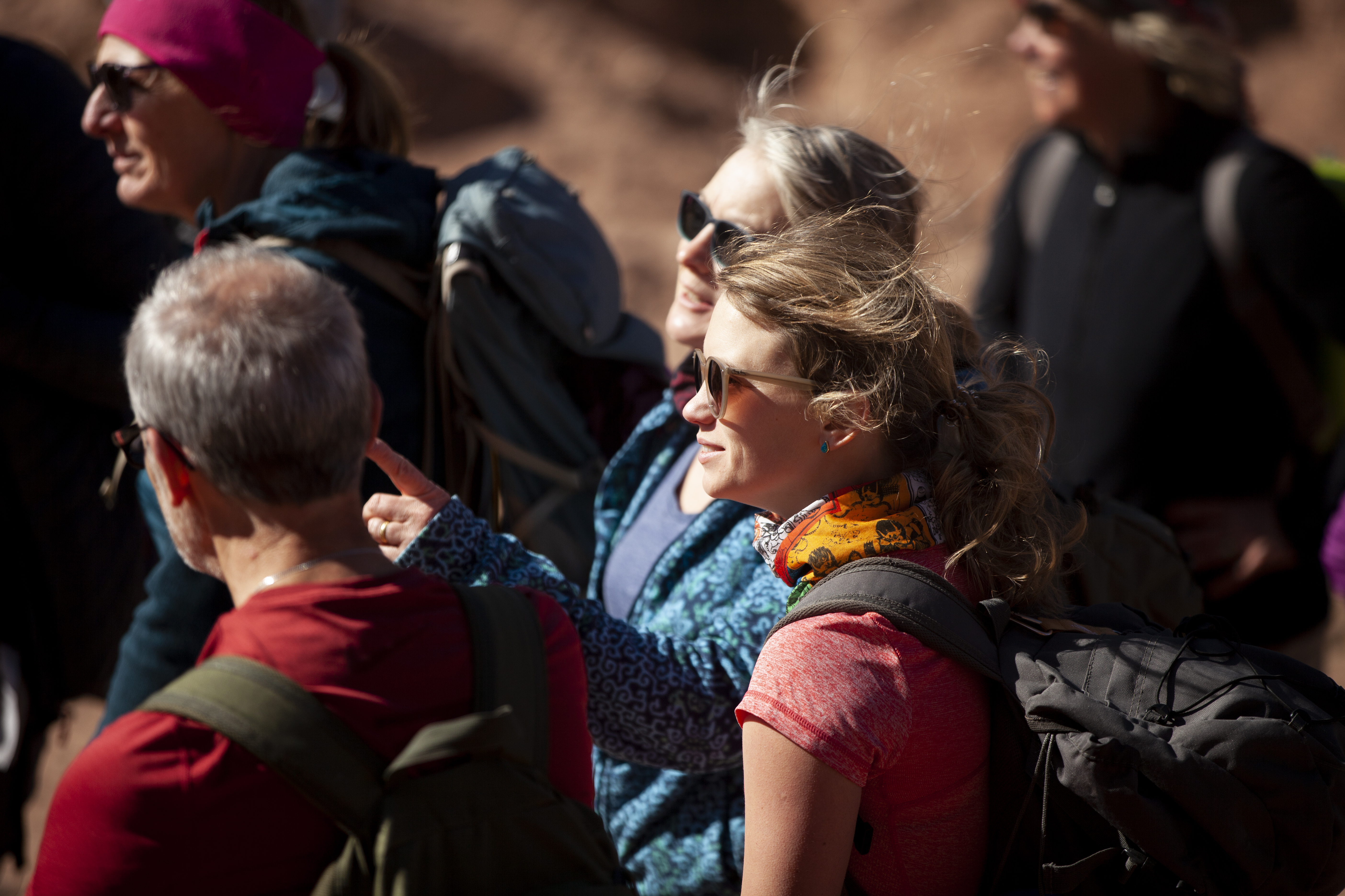 A group of hikers, wearing backpacks and sunglasses, stand together in sunlight, conversing and pointing, in a desert-like environment. They appear engaged and ready for adventure.