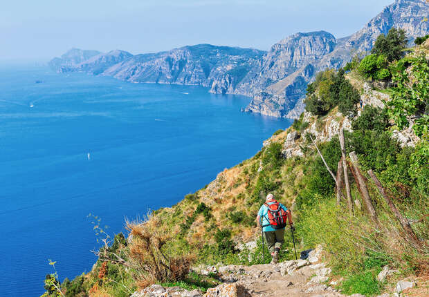 A hiker with a red backpack walks down a rocky trail along a cliff, overlooking a vast blue sea and distant mountainous coastline under a clear sky.