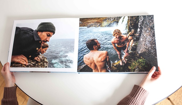 A person holds an open photo book. Left page shows two people in cold weather attire looking out over the ocean. Right page depicts three shirtless individuals near a cliff waterfall.