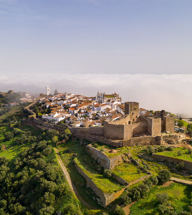 A historic castle crowns a hilltop village, with white houses and a church, surrounded by fortifications. Lush greenery covers the landscape, and clouds encircle the distant horizon.