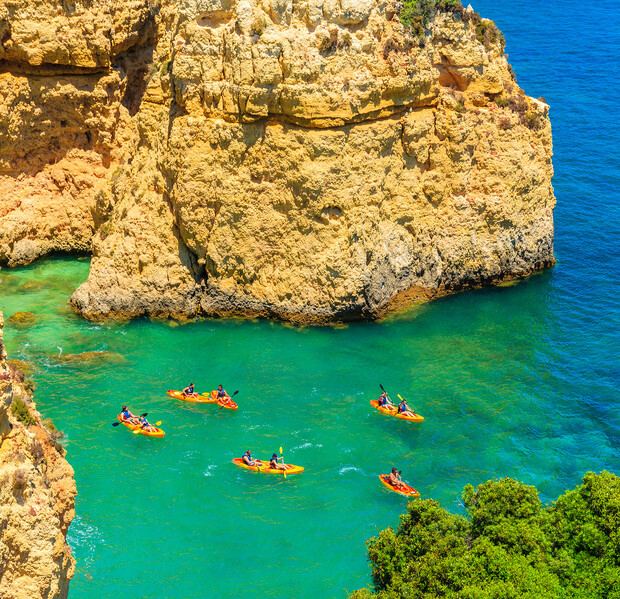 Kayakers paddling in bright orange kayaks on turquoise water beside large, rugged cliffs, surrounded by lush greenery under a clear, sunny sky.
