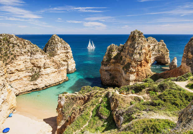 A sailboat glides across a vibrant blue ocean, surrounded by rugged cliffs and a secluded sandy beach under a clear, blue sky.
