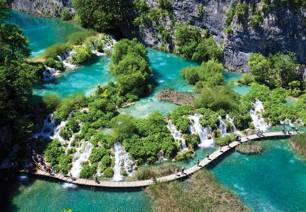 Turquoise waterfalls cascade through lush greenery, as people walk along a winding wooden boardwalk, surrounded by vibrant pools and rocky cliffs in a serene natural setting.