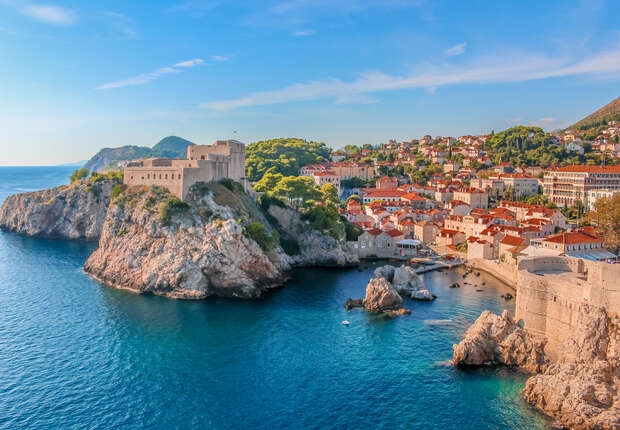 A historic fortress stands on a rocky cliff, overlooking a vibrant coastal town with terracotta rooftops and lush greenery, adjacent to a deep blue sea under a clear sky.
