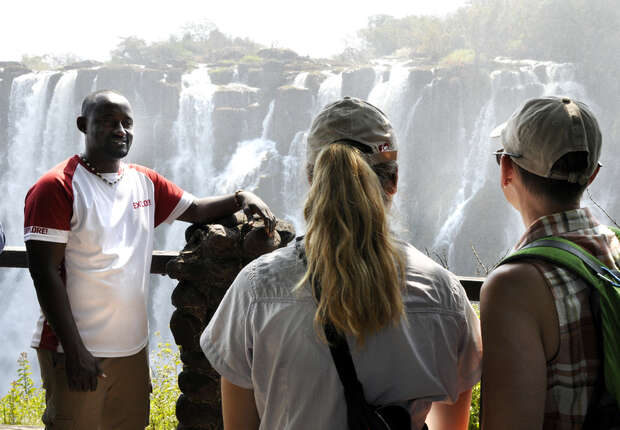 A person leans against a stone railing, speaking to two others facing them, with a large waterfall cascading in the background. Bright daylight filters through the lush surroundings.